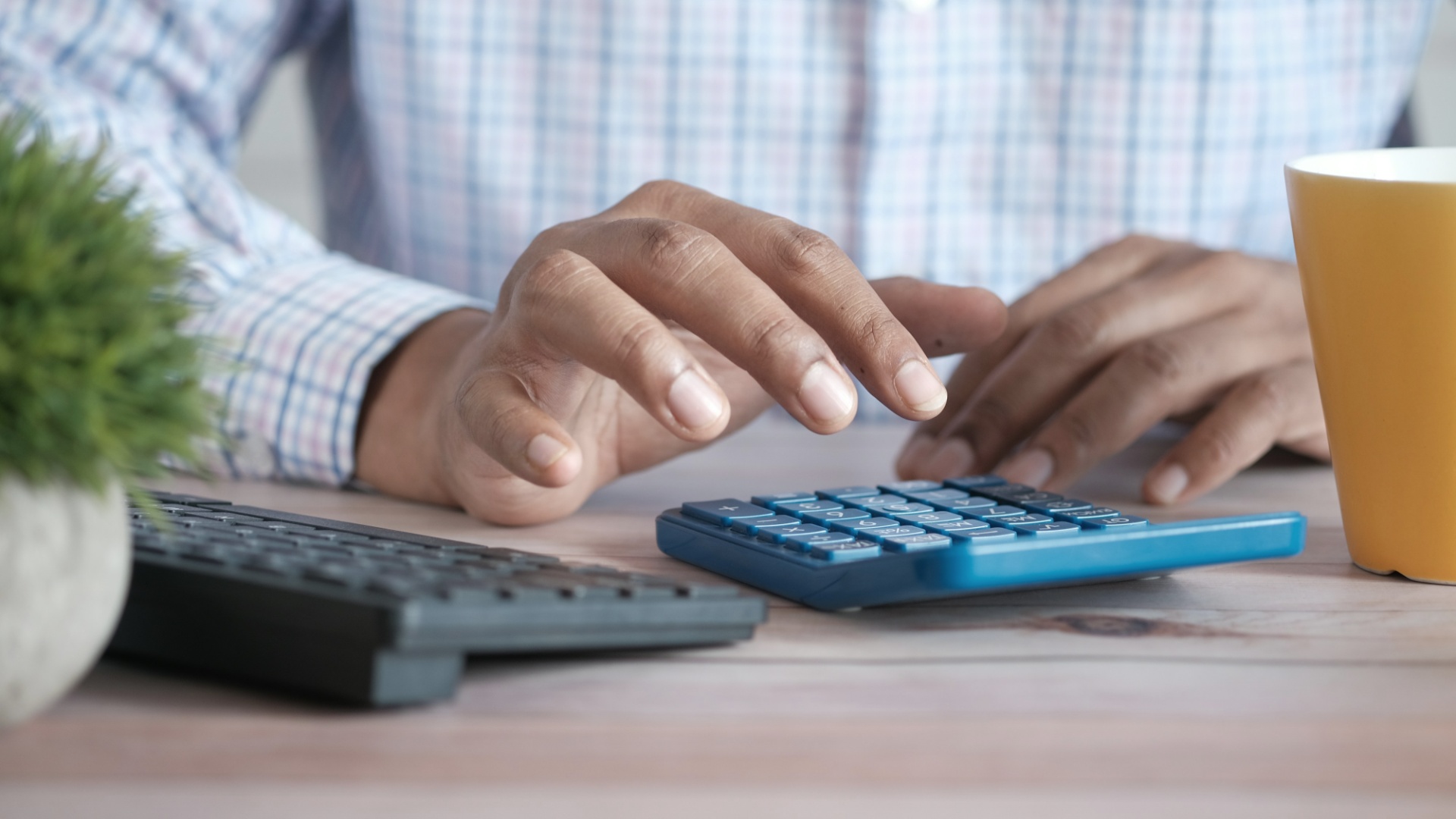 A person's hands pressing the keys of a blue calculator on a wooden desk, with a computer keyboard, a small plant and a yellow mug beside it.