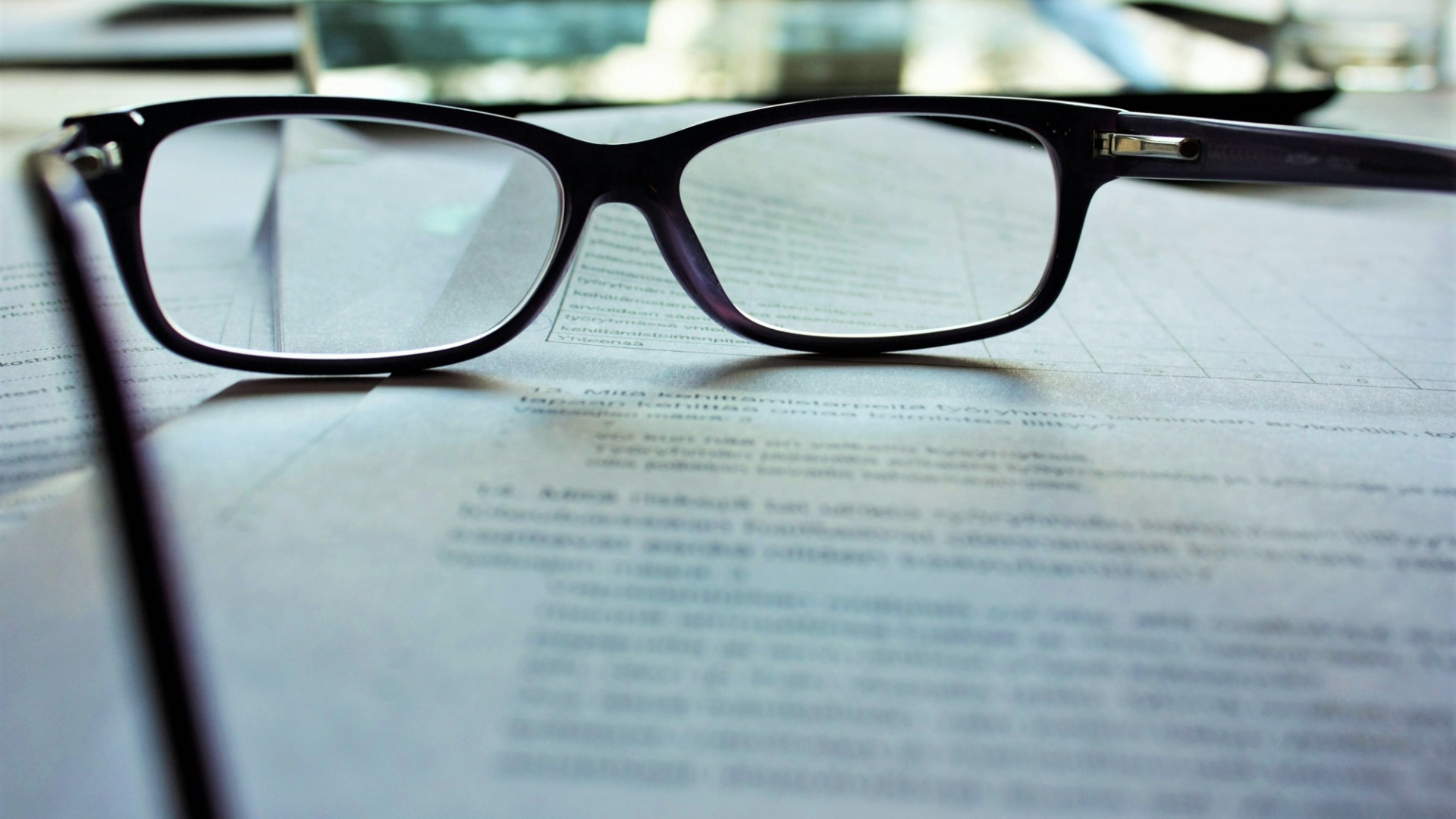 A pair of black-framed reading glasses resting on printed paper forms, with the page text out of focus.