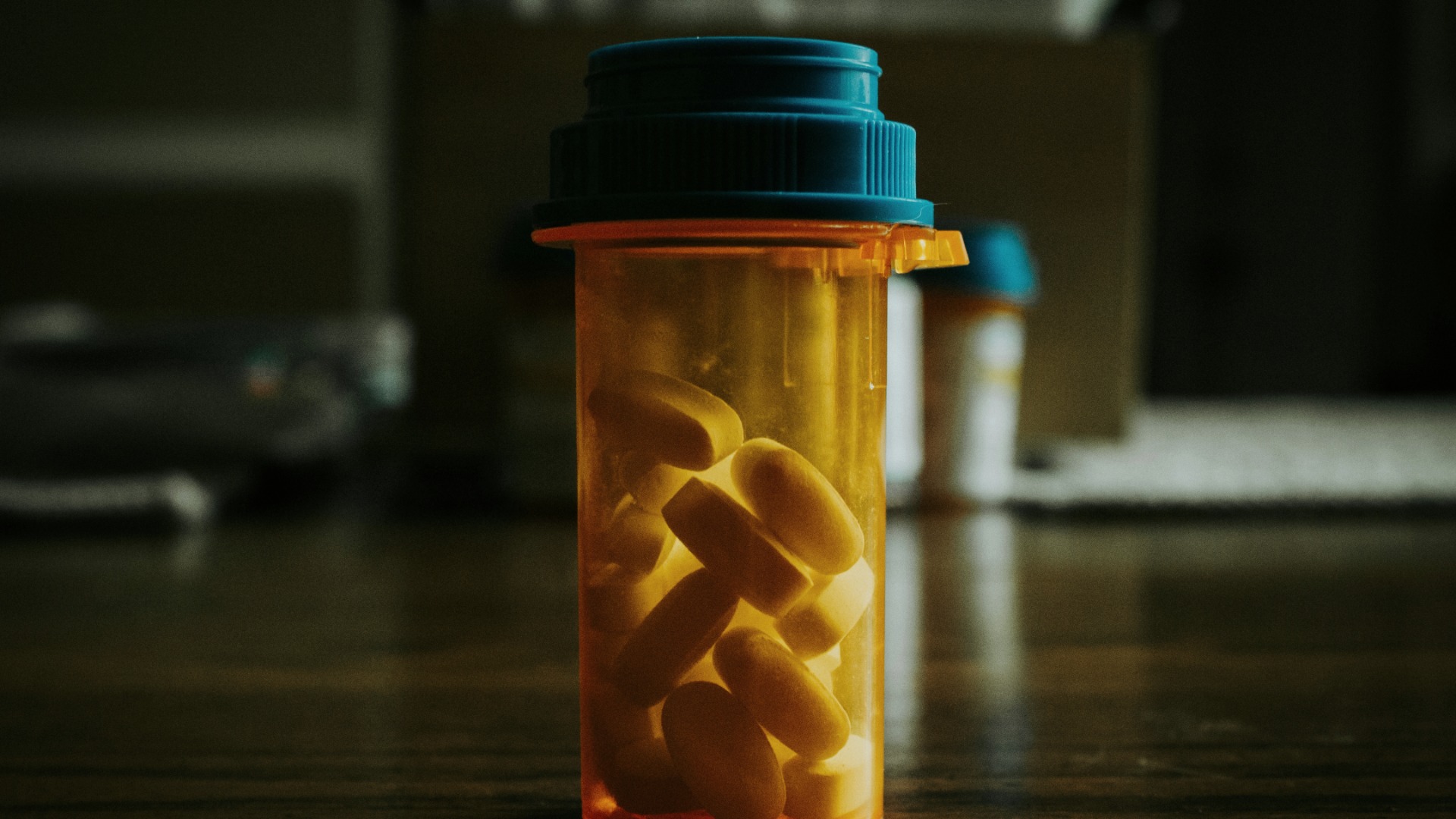 A single amber prescription pill bottle with a blue cap, filled with oval white pills, standing on a dark wooden table.