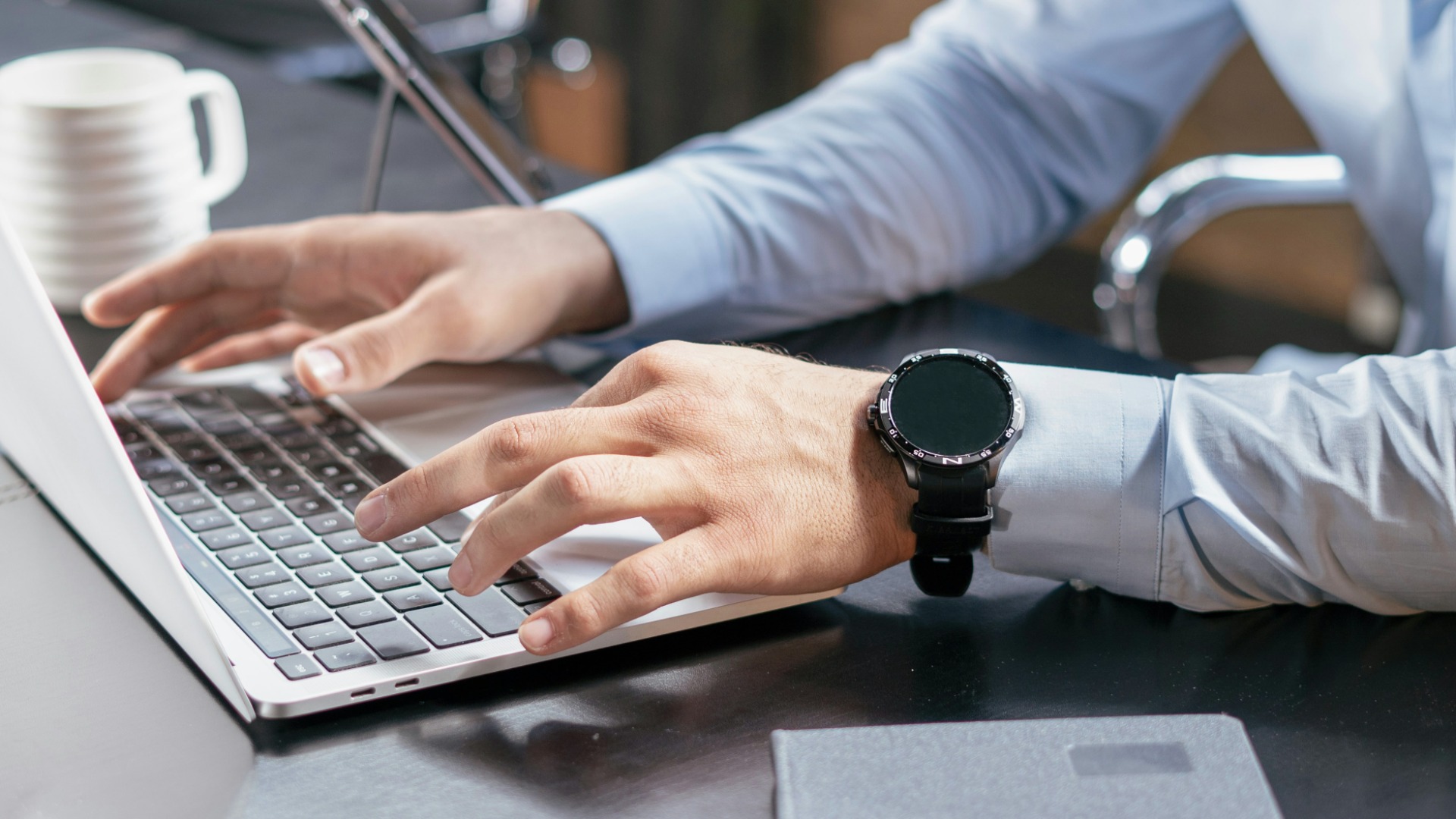 Two hands typing on a silver laptop at a dark desk, with a white coffee mug and a tablet on a stand in the background.