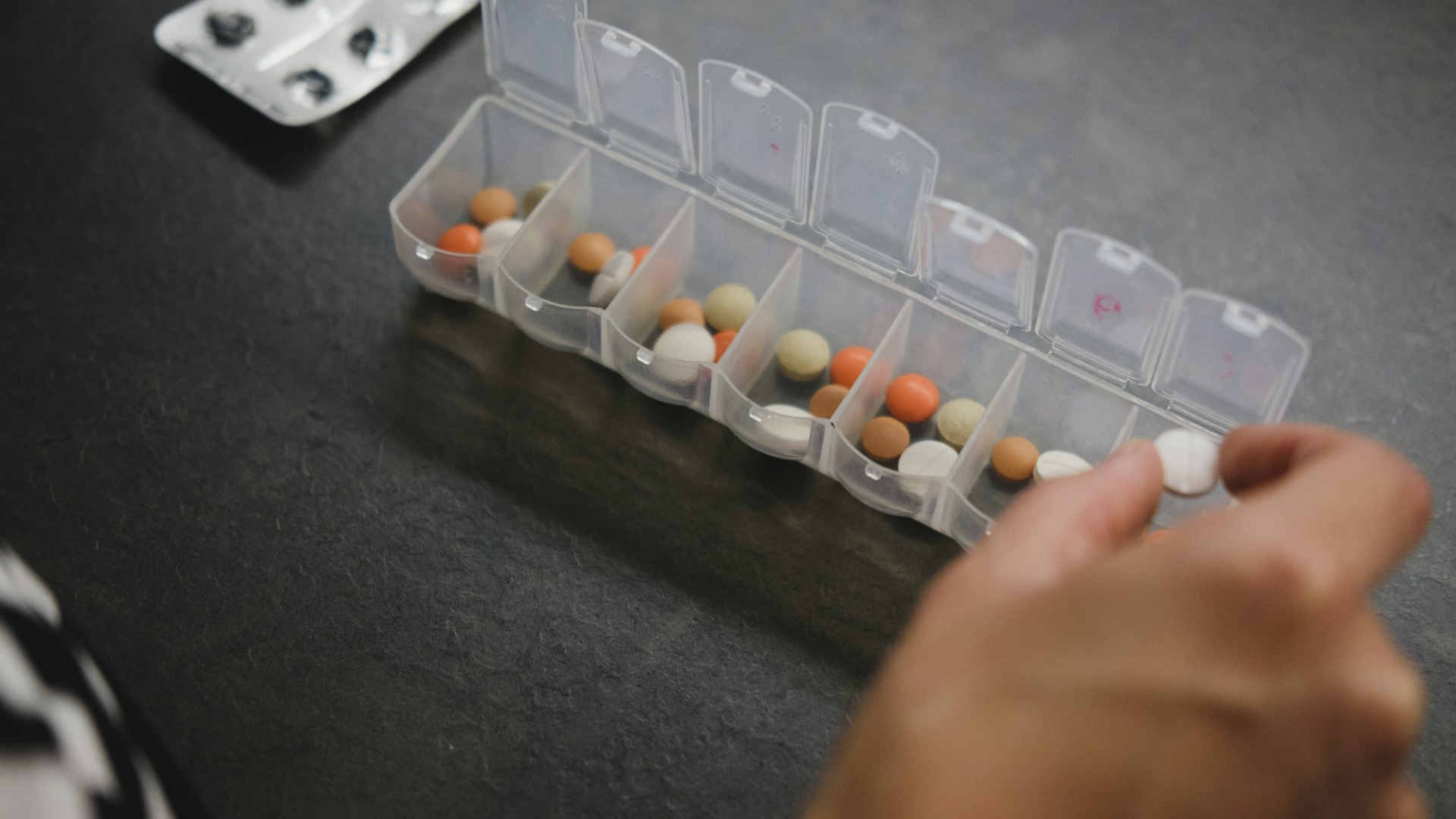 A hand reaching for a white pill in a clear plastic weekly pill organizer holding orange, white and tan pills, on a dark slate surface.